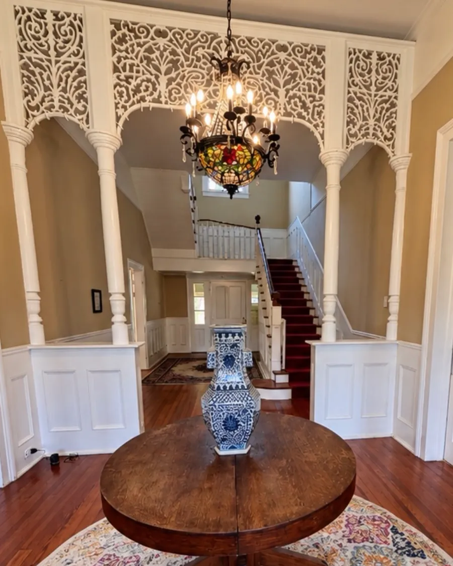 The entry hall at VanCleave Hall: a white fretwork arch on turned columns and a stained-glass chandelier above a round table, with the red-carpeted staircase beyond