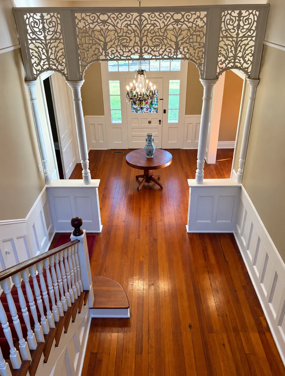 Looking down from the stair landing into the entry hall: a white fretwork arch, a crystal chandelier before the stained-glass front door, and a round table on heart-pine floors